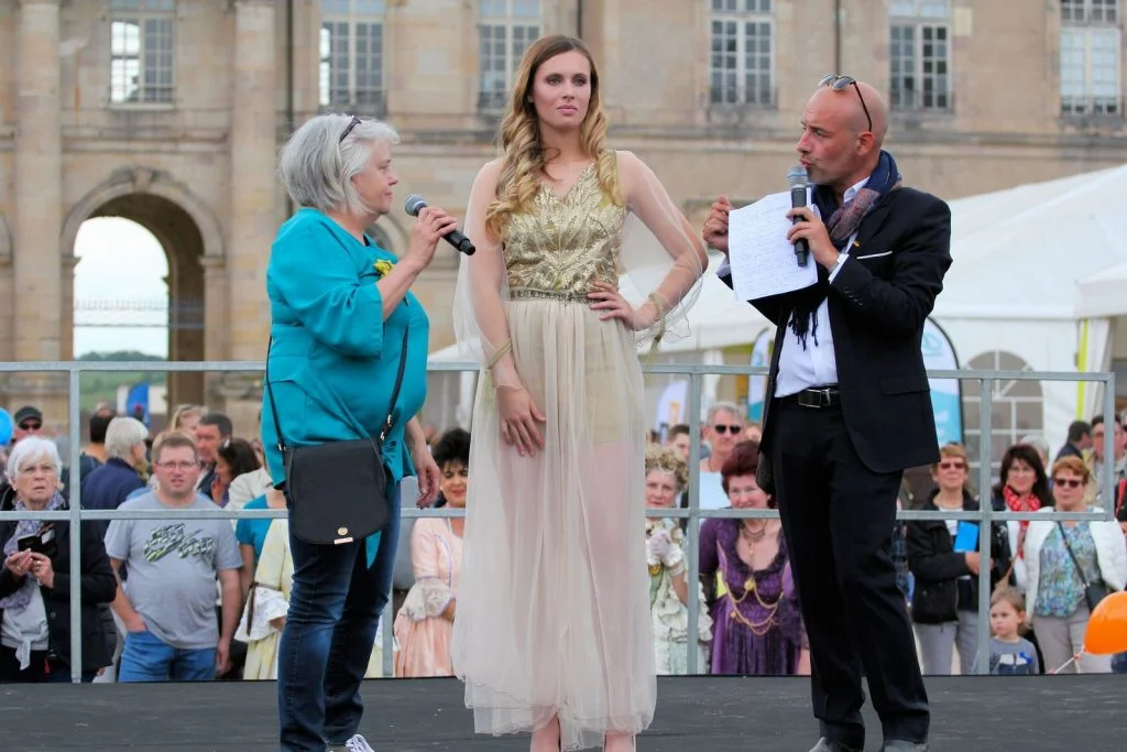 presentation sur un podium d'une collection de mode de brimbel et mirgamote creatrice de mode, avec un modèle portant une robe couleur champagne et un présentateur présentant la créatrice et la tenue phare de la collection