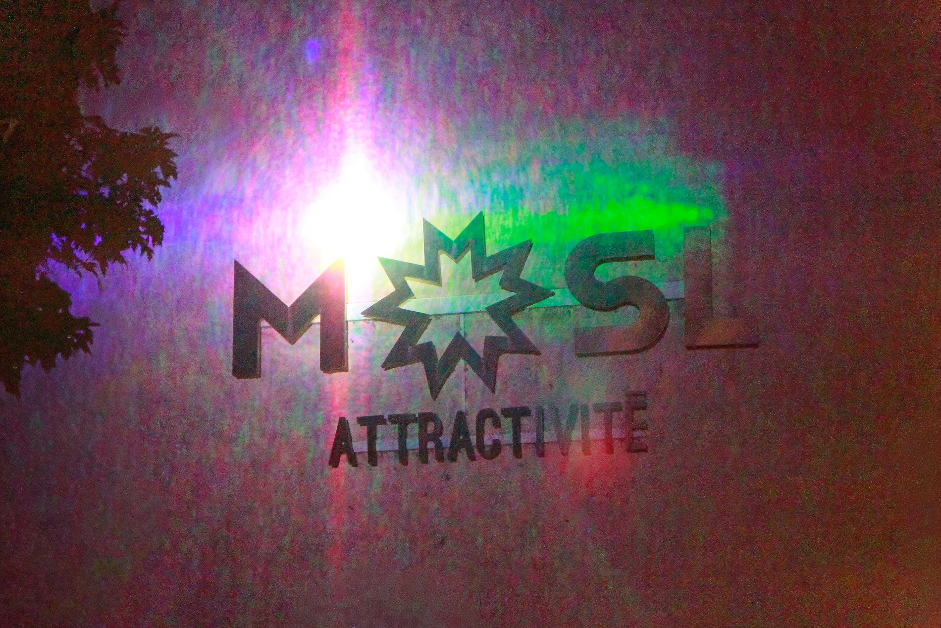 logo lumineux multicolore de mosl agence d attractivite, implanté sur le mur d'un immeuble pres d un pont, dans le cadre de la manifestation constellations metz 2026